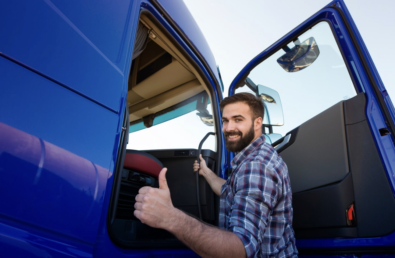 A professional truck driver standing next to a large truck, giving a thumbs-up sign.